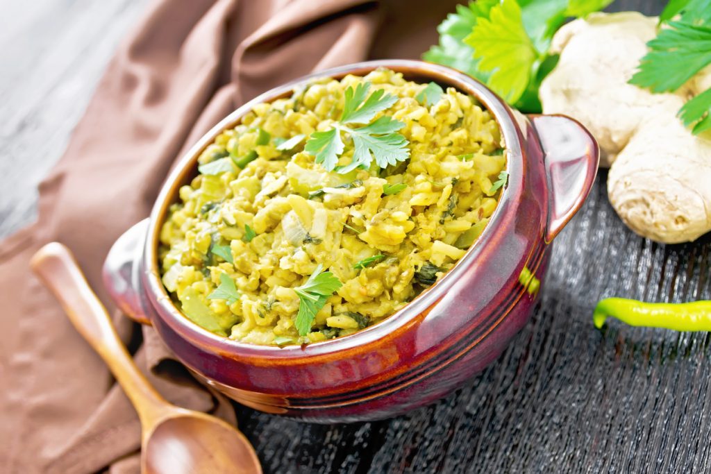 Indian national dish of kichari made of mung bean, rice, celery, spinach, hot pepper and spices in a bowl on a napkin, ginger and spoon on dark wooden board background